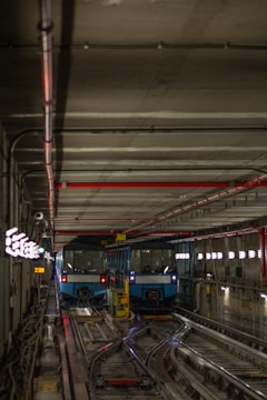Two blue metro trains are parked side by side on separate tracks inside a dimly lit underground subway tunnel. The tunnel features a concrete ceiling with red and black piping, illuminated directional signs on the left, and glowing lights on the right wall. Tracks and various technical installations are visible on the floor.