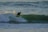 A surfer in a wetsuit is riding a wave in the ocean, skillfully balancing on the surfboard as the water splashes around them. The scene captures the dynamic motion and energy of surfing in an expansive seascape.