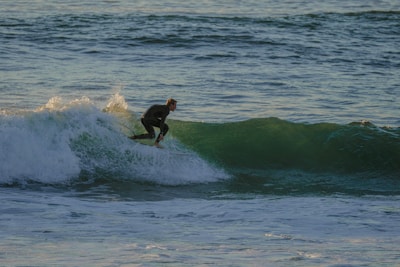 A surfer in a wetsuit is riding a wave in the ocean, skillfully balancing on the surfboard as the water splashes around them. The scene captures the dynamic motion and energy of surfing in an expansive seascape.