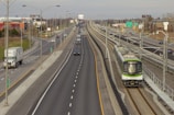 A highway scene with multiple lanes of traffic, including cars and a large truck, running parallel to a train track. A modern green and white train is approaching from the distance on the right side. Overhead power lines and street lamps are visible, along with signs, some greenery, and commercial buildings in the background.