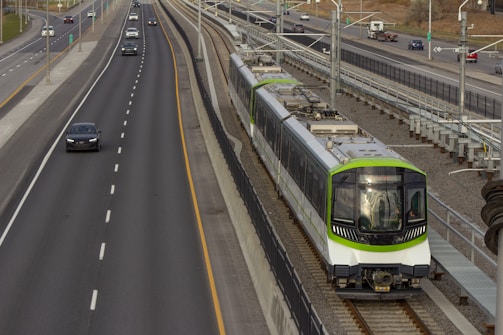 A modern light rail train travels on a dedicated track parallel to a multi-lane highway. Several cars are visible on the highway, and the surrounding area appears urban with a mix of road infrastructure and some greenery in the background. The train features a green and white color scheme.