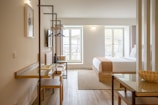 A modern, well-lit hotel room with wooden flooring and contemporary furnishings. There is a neatly made bed with white linens and a beige throw, a desk with a chair, a wall-mounted TV, and a large window providing natural light. The room features clean lines and minimalistic decor.