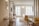 A modern, well-lit hotel room with wooden flooring and contemporary furnishings. There is a neatly made bed with white linens and a beige throw, a desk with a chair, a wall-mounted TV, and a large window providing natural light. The room features clean lines and minimalistic decor.