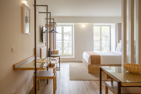A modern, well-lit hotel room with wooden flooring and contemporary furnishings. There is a neatly made bed with white linens and a beige throw, a desk with a chair, a wall-mounted TV, and a large window providing natural light. The room features clean lines and minimalistic decor.