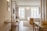 A modern, well-lit hotel room with wooden flooring and contemporary furnishings. There is a neatly made bed with white linens and a beige throw, a desk with a chair, a wall-mounted TV, and a large window providing natural light. The room features clean lines and minimalistic decor.