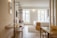 A modern, well-lit hotel room with wooden flooring and contemporary furnishings. There is a neatly made bed with white linens and a beige throw, a desk with a chair, a wall-mounted TV, and a large window providing natural light. The room features clean lines and minimalistic decor.
