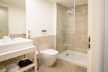 A modern bathroom featuring a glass-enclosed shower area, a wall-mounted toilet, and a sleek rectangular sink on a vanity with storage space underneath. The walls and floor are tiled in a light beige color, and there is a towel and toiletries visible on the vanity shelf.
