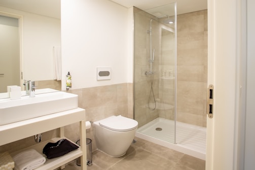 A modern bathroom featuring a glass-enclosed shower area, a wall-mounted toilet, and a sleek rectangular sink on a vanity with storage space underneath. The walls and floor are tiled in a light beige color, and there is a towel and toiletries visible on the vanity shelf.