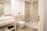 A modern bathroom featuring a glass-enclosed shower area, a wall-mounted toilet, and a sleek rectangular sink on a vanity with storage space underneath. The walls and floor are tiled in a light beige color, and there is a towel and toiletries visible on the vanity shelf.