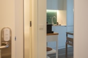 A modern kitchen area is partially visible through an open door. The design features white cabinetry, a green tiled backsplash, and a sleek faucet. A small table and chair are situated nearby, with neutral tones creating a minimalist aesthetic. The lighting is warm, contributing to a cozy ambiance.