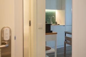 A modern kitchen area is partially visible through an open door. The design features white cabinetry, a green tiled backsplash, and a sleek faucet. A small table and chair are situated nearby, with neutral tones creating a minimalist aesthetic. The lighting is warm, contributing to a cozy ambiance.