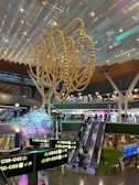 An interior view of a modern airport terminal featuring a large, intricate, gold sculpture hanging from the ceiling. Below the sculpture, several travelers can be seen moving through the area. There are multiple illuminated signs displaying gate information in both English and Arabic. Escalators lead to an upper level where shops and eateries are visible. The space is well-lit, with various colors highlighting the structures and decorations.