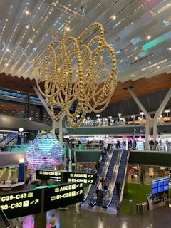 An interior view of a modern airport terminal featuring a large, intricate, gold sculpture hanging from the ceiling. Below the sculpture, several travelers can be seen moving through the area. There are multiple illuminated signs displaying gate information in both English and Arabic. Escalators lead to an upper level where shops and eateries are visible. The space is well-lit, with various colors highlighting the structures and decorations.