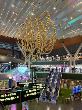 An interior view of a modern airport terminal featuring a large, intricate, gold sculpture hanging from the ceiling. Below the sculpture, several travelers can be seen moving through the area. There are multiple illuminated signs displaying gate information in both English and Arabic. Escalators lead to an upper level where shops and eateries are visible. The space is well-lit, with various colors highlighting the structures and decorations.