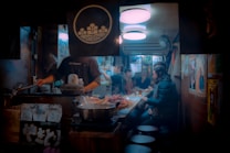 An indoor restaurant scene with a chef cooking on the left side and people seated at a counter on the right. The counter is laden with food items and there are various posters and decorations on the walls. The lighting is warm and intimate, creating a cozy atmosphere. Steam can be seen rising from pots, adding to the bustling and warm ambiance.