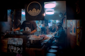 An indoor restaurant scene with a chef cooking on the left side and people seated at a counter on the right. The counter is laden with food items and there are various posters and decorations on the walls. The lighting is warm and intimate, creating a cozy atmosphere. Steam can be seen rising from pots, adding to the bustling and warm ambiance.