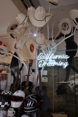 A display features a collection of white cowboy hats hanging on white branches, three acoustic guitars leaning against a wall, and a neon sign reading 'California Dreaming'. Below are several motorcycle helmets in various designs. The background includes a poster of a horse and a person partly visible to the side.
