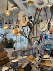 A store interior features a display with several hats hanging on branches, illuminated by warm hanging lightbulbs. The surrounding area is decorated with various homeware products including patterned dishes, glassware, and rolled-up textiles. A straw basket, a stuffed animal, and framed artwork add to the eclectic arrangement. The setting suggests a cozy and rustic ambiance.