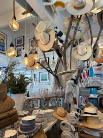 A store interior features a display with several hats hanging on branches, illuminated by warm hanging lightbulbs. The surrounding area is decorated with various homeware products including patterned dishes, glassware, and rolled-up textiles. A straw basket, a stuffed animal, and framed artwork add to the eclectic arrangement. The setting suggests a cozy and rustic ambiance.