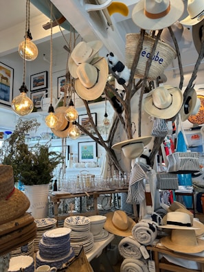 A store interior features a display with several hats hanging on branches, illuminated by warm hanging lightbulbs. The surrounding area is decorated with various homeware products including patterned dishes, glassware, and rolled-up textiles. A straw basket, a stuffed animal, and framed artwork add to the eclectic arrangement. The setting suggests a cozy and rustic ambiance.