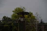 A metal structure with an old wheel on top, likely part of an industrial or electrical installation, is set against the backdrop of trees and a cloudy sky. Several power lines run horizontally across the image.