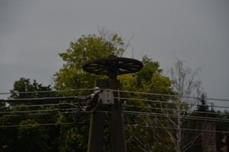A metal structure with an old wheel on top, likely part of an industrial or electrical installation, is set against the backdrop of trees and a cloudy sky. Several power lines run horizontally across the image.