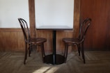Two wooden chairs are placed on either side of a small, dark-brown wooden table. The setting is in a room with wooden wall panels and beige carpeted floor.