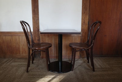 Two wooden chairs are placed on either side of a small, dark-brown wooden table. The setting is in a room with wooden wall panels and beige carpeted floor.