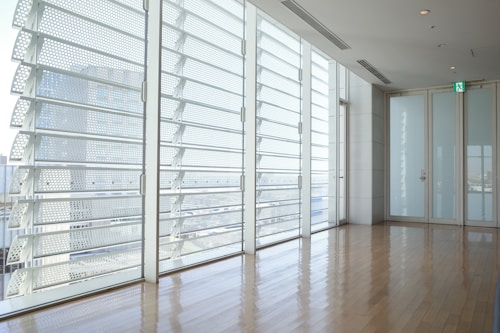 A modern interior space featuring large floor-to-ceiling windows with horizontal blinds. The room has polished wooden floors that reflect light, and there is a glass door with an emergency exit sign above it. The exterior view includes blurred skyscrapers, suggesting an urban environment.