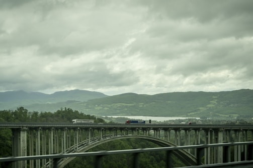 A convoy of metallic trucks crossing a modern bridge under a silver sky.