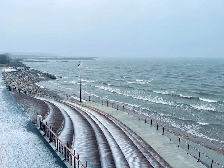 A coastal scene featuring a wooden amphitheater with curved seating facing a rough sea. The overcast sky and gray waves create a cold and windy atmosphere. The shore is lined with rocks and a lone streetlamp stands along the pathway.