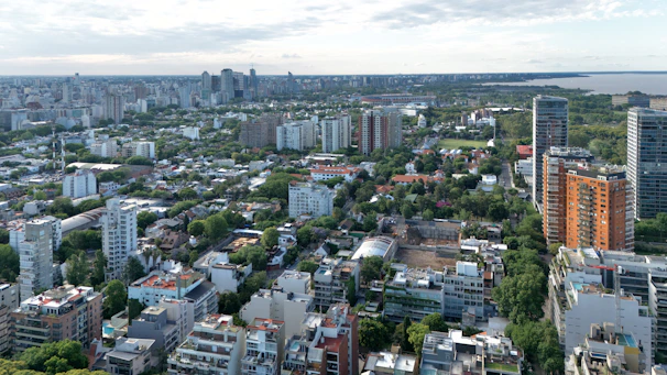 An aerial view of a cityscape with a mix of residential and commercial buildings surrounded by greenery. High-rise buildings are scattered across the scene, with mid-rise and low-rise structures filling in the gaps. The landscape stretches towards a distant body of water, with a cloudy sky overhead.