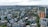 An aerial view of a cityscape with a mix of residential and commercial buildings surrounded by greenery. High-rise buildings are scattered across the scene, with mid-rise and low-rise structures filling in the gaps. The landscape stretches towards a distant body of water, with a cloudy sky overhead.