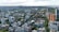 An aerial view of a cityscape with a mix of residential and commercial buildings surrounded by greenery. High-rise buildings are scattered across the scene, with mid-rise and low-rise structures filling in the gaps. The landscape stretches towards a distant body of water, with a cloudy sky overhead.