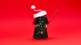 A small figurine of a character dressed in black armor, holding a red lightsaber, and wearing a red and white Santa hat, set against a solid red background.