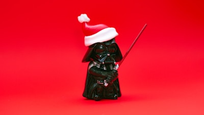 A small figurine of a character dressed in black armor, holding a red lightsaber, and wearing a red and white Santa hat, set against a solid red background.