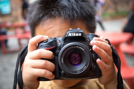 A child holds a Nikon camera up to their face, preparing to take a photo. The camera is centrally positioned, with a focus on the lens. The background appears blurred, including red benches.