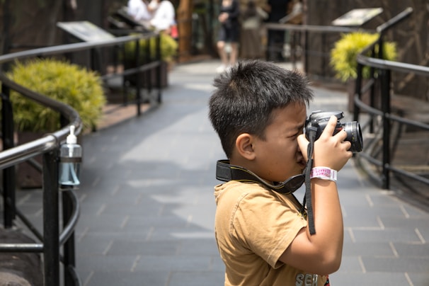 A young boy is holding a camera to his face, using it to take a photo. He is wearing a tan shirt and has a wristband on his left wrist. His short hair gives him a neat appearance and he is standing on a tiled path outdoors, surrounded by metal railings and greenery.