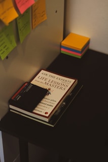A desk setup featuring office supplies such as colorful sticky notes, a book titled 'Life Lessons from the Masters,' a pen, and a closed black notebook. The scene conveys a workspace environment with a focus on organization and productivity.
