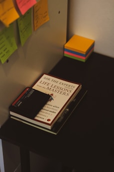 A desk setup featuring office supplies such as colorful sticky notes, a book titled 'Life Lessons from the Masters,' a pen, and a closed black notebook. The scene conveys a workspace environment with a focus on organization and productivity.