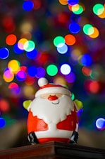 A cheerful ceramic Santa Claus figurine is positioned in front of a vibrant and blurred background of colorful bokeh lights, creating a festive and joyful atmosphere.