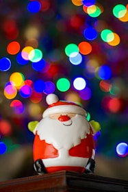 A cheerful ceramic Santa Claus figurine is positioned in front of a vibrant and blurred background of colorful bokeh lights, creating a festive and joyful atmosphere.