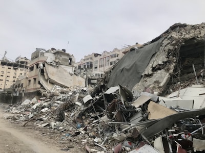 A scene of extensive urban destruction with collapsed buildings and piles of debris. The remains of the structures are comprised of concrete, metal, and various building materials. The environment appears desolate and the sky is overcast.