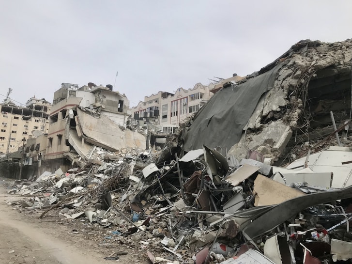 A scene of extensive urban destruction with collapsed buildings and piles of debris. The remains of the structures are comprised of concrete, metal, and various building materials. The environment appears desolate and the sky is overcast.