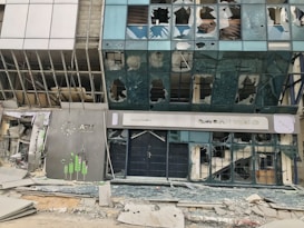 A building with extensive damage, with shattered glass windows and debris scattered on the ground. The facade is severely impacted, with signs for an ATM and a bank visible but also damaged.