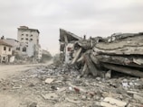 Several buildings are damaged and partially collapsed, with rubble and debris scattered across the ground. The sky is overcast, and there is a deserted street leading into the distance. The scene appears desolate and chaotic, suggesting recent destruction.