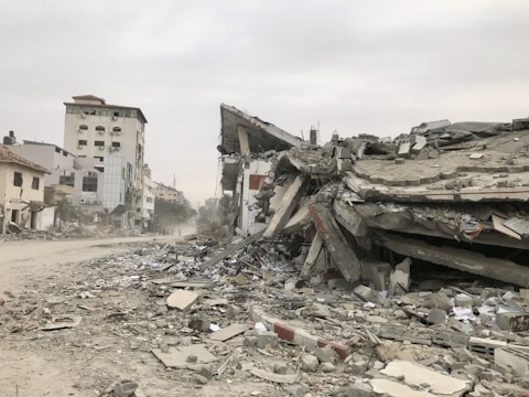 Several buildings are damaged and partially collapsed, with rubble and debris scattered across the ground. The sky is overcast, and there is a deserted street leading into the distance. The scene appears desolate and chaotic, suggesting recent destruction.