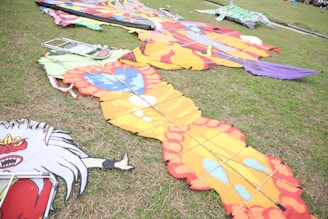Vibrantly colored kites with intricate designs are laid out on a grassy field. One has an orange and yellow pattern with red edges, while another is decorated with a fierce red face. A metal folding chair is placed near the kites, and additional colorful kite parts are visible in the background.