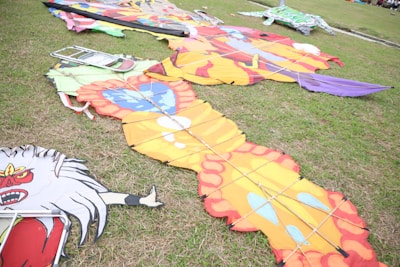 Vibrantly colored kites with intricate designs are laid out on a grassy field. One has an orange and yellow pattern with red edges, while another is decorated with a fierce red face. A metal folding chair is placed near the kites, and additional colorful kite parts are visible in the background.