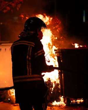 A firefighter in protective gear stands in front of a large, outdoor fire, holding a hose. The flames illuminate the scene, creating an intense and urgent atmosphere.